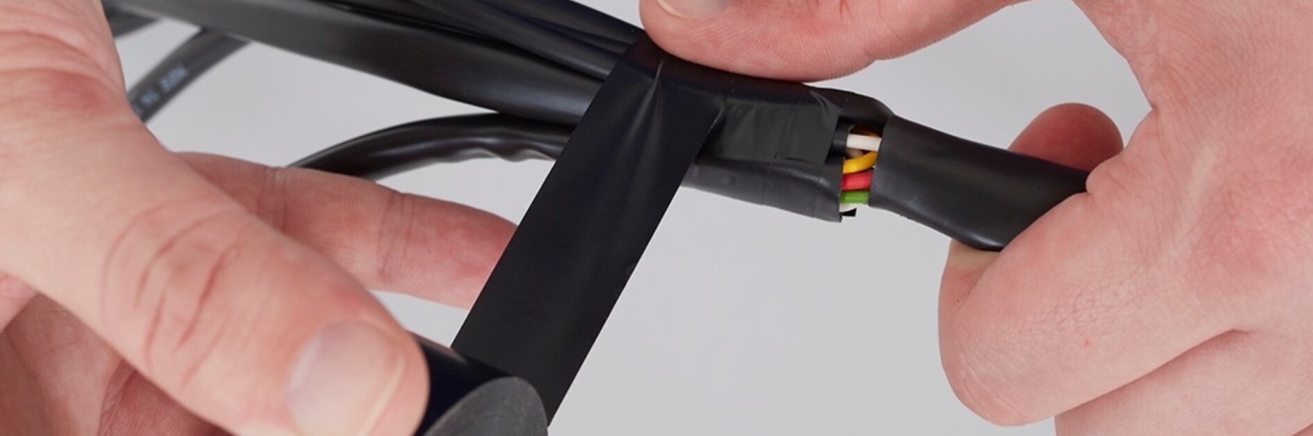 A person wraps black tesa tape around a group of colored wires. The tesa tape is being applied to secure and insulate the wires, which include green, yellow, and red colors. The background is plain white. (This text has been generated by AI)