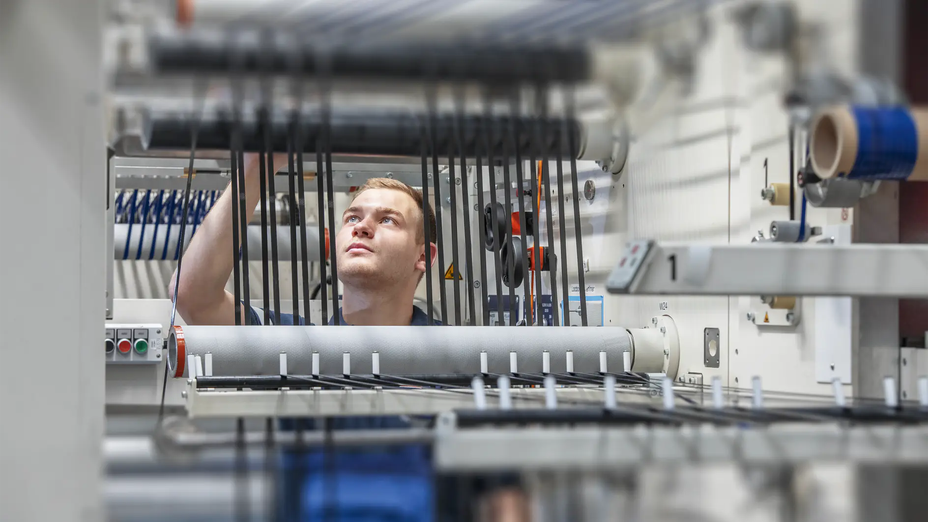 A person in a blue uniform works with machinery in a factory setting, adjusting black cables on a series of rollers and levers using tesa tape. The environment is industrial, with various mechanical components and controls visible.