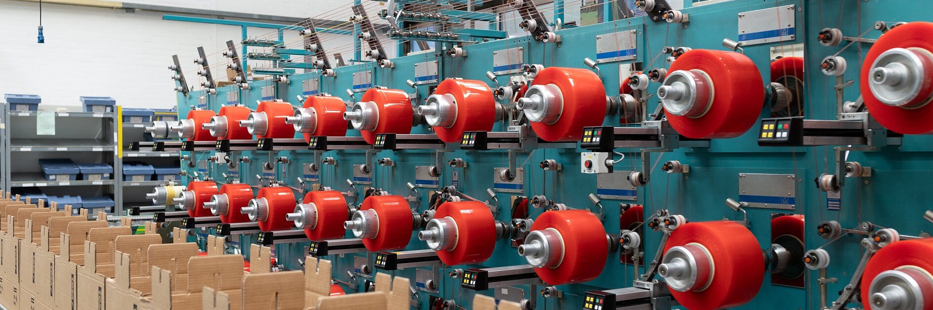 Factory production line featuring teal machines, red thread spools, rolls of tesa tape, and cardboard boxes on a conveyor belt, showcasing industrial adhesives solutions for efficient manufacturing and packaging operations.