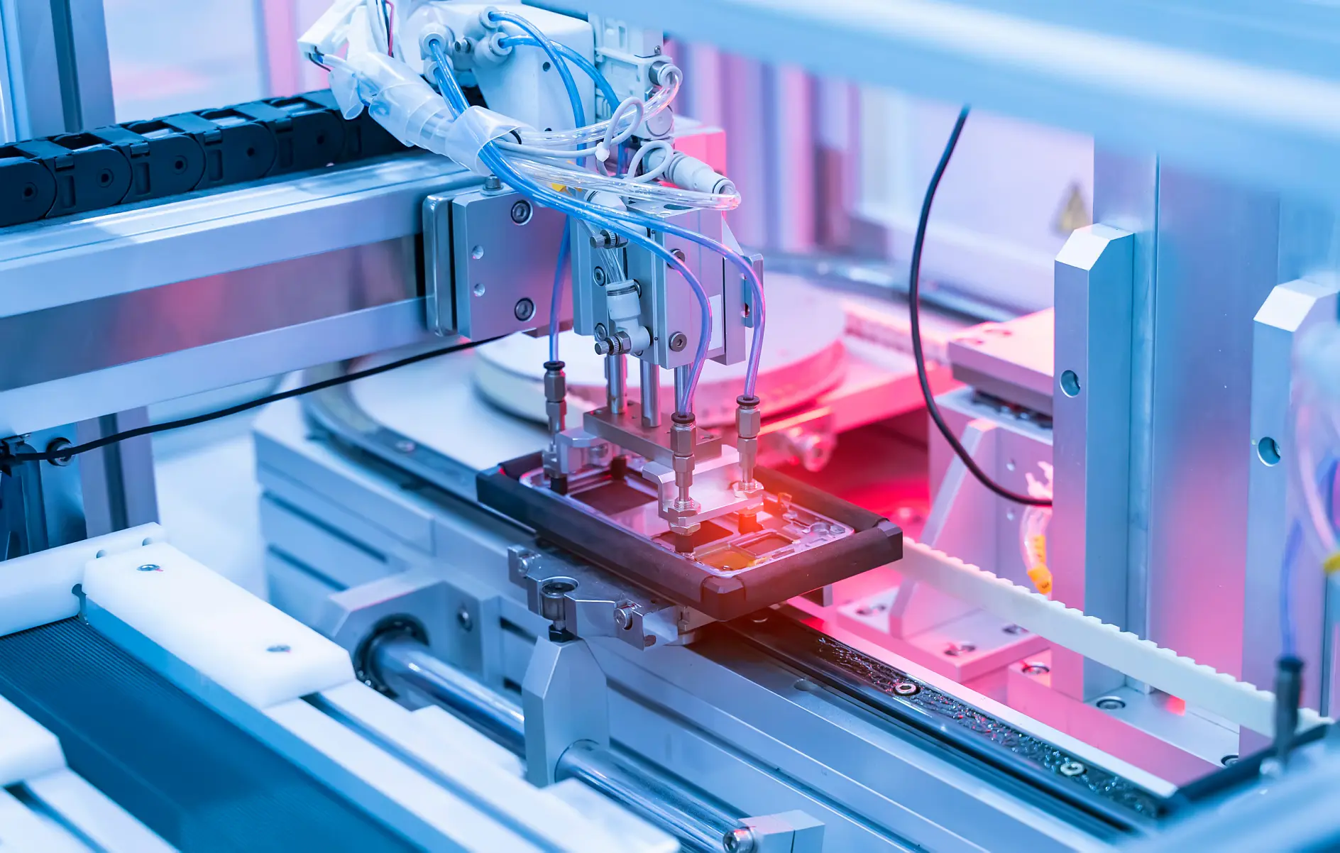 A robotic arm operates on an electronic device in a manufacturing facility, highlighted with pink and blue lights, showcasing precision machinery used in automated production processes. Various cables, mechanical parts, and rolls of tesa electronic adhesive foam tapes are visible. (This text has been generated by AI)