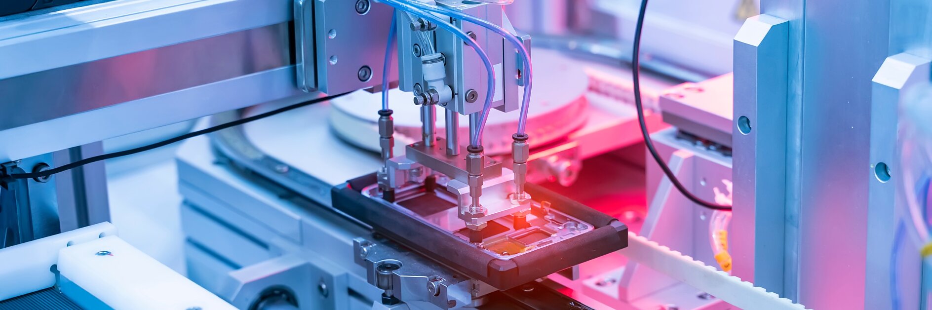 A robotic arm operates on an electronic device in a manufacturing facility, highlighted with pink and blue lights, showcasing precision machinery used in automated production processes. Various cables, mechanical parts, and rolls of tesa electronic adhesive foam tapes are visible.