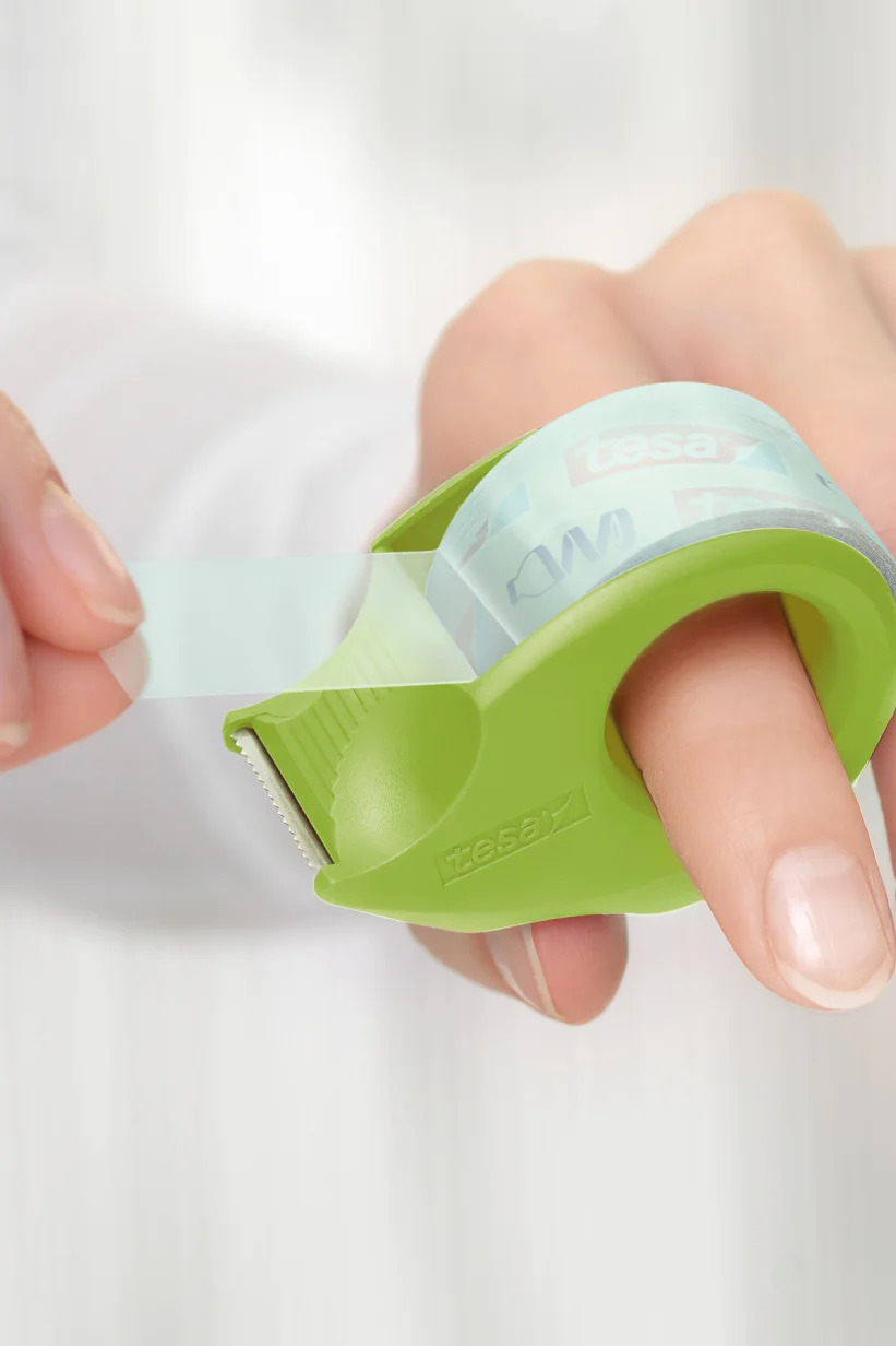 tesafilm-mini-dispenser-eco-crystal_model_close-up_ap-Print (2) Close-up of a person dispensing clear adhesive tesa tape using a green handheld tape dispenser. The dispenser is wrapped around the index finger, and the person is pulling the tesa tape with the other hand. The background is softly blurred. (This text has been generated by AI)
