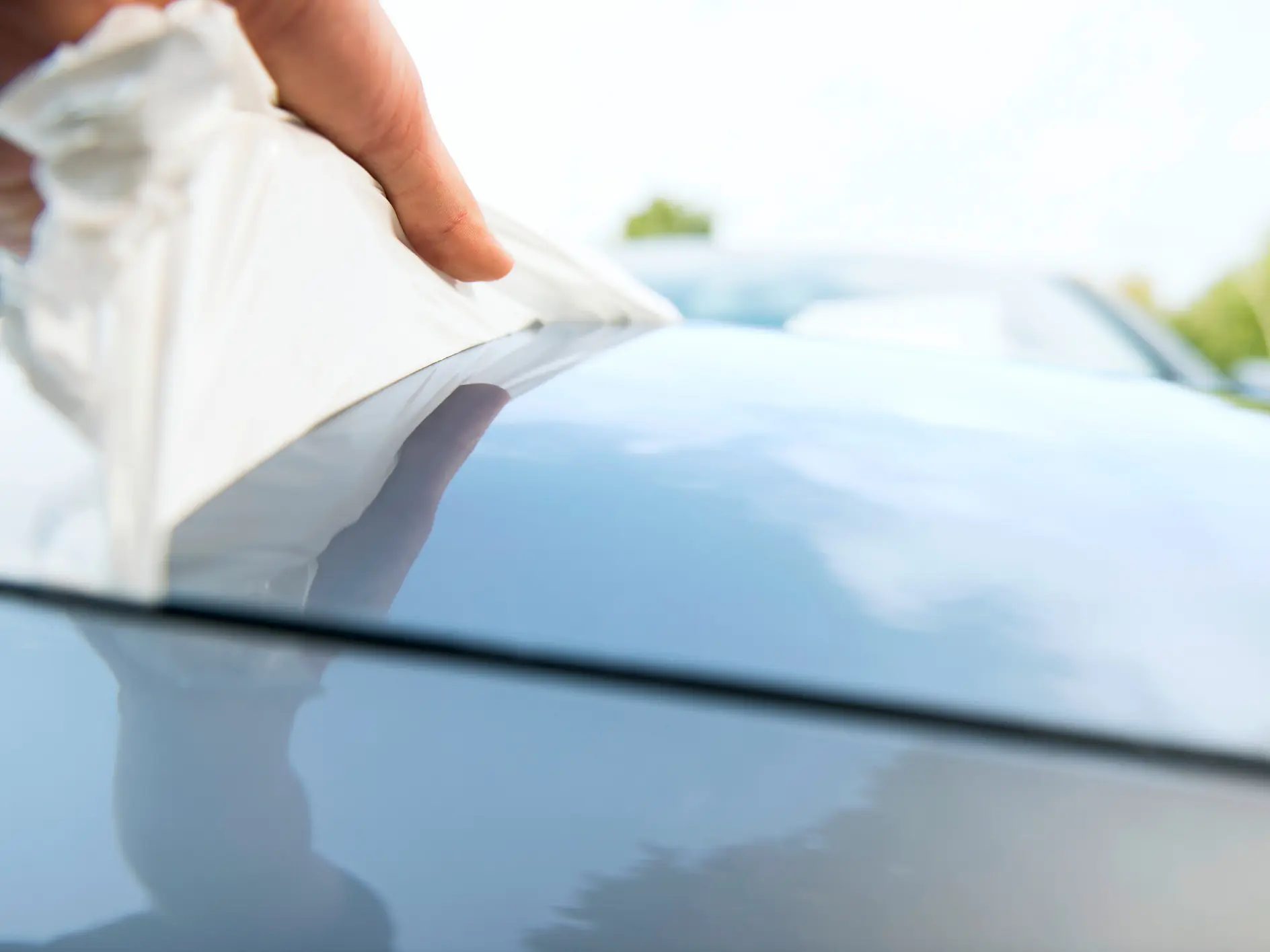 hand using a white tesa cloth to clean the shiny surface of a car hood outdoors on a bright day (This text has been generated by AI)