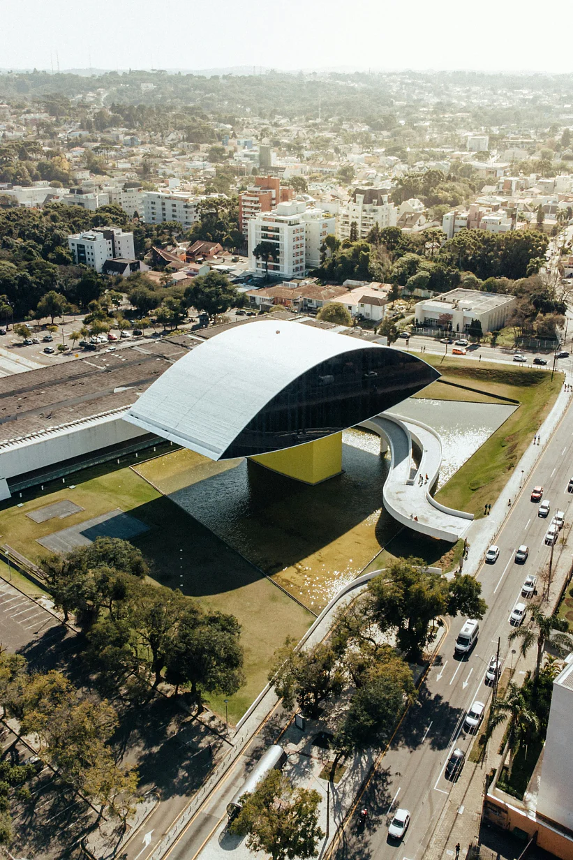 Aerial view of the Oscar Niemeyer Museum in Curitiba, Brazil. The building features a distinctive eye-shaped structure with a reflective facade, surrounded by a green lawn. Roads with traffic and surrounding residential areas are visible. (This text has been generated by AI)