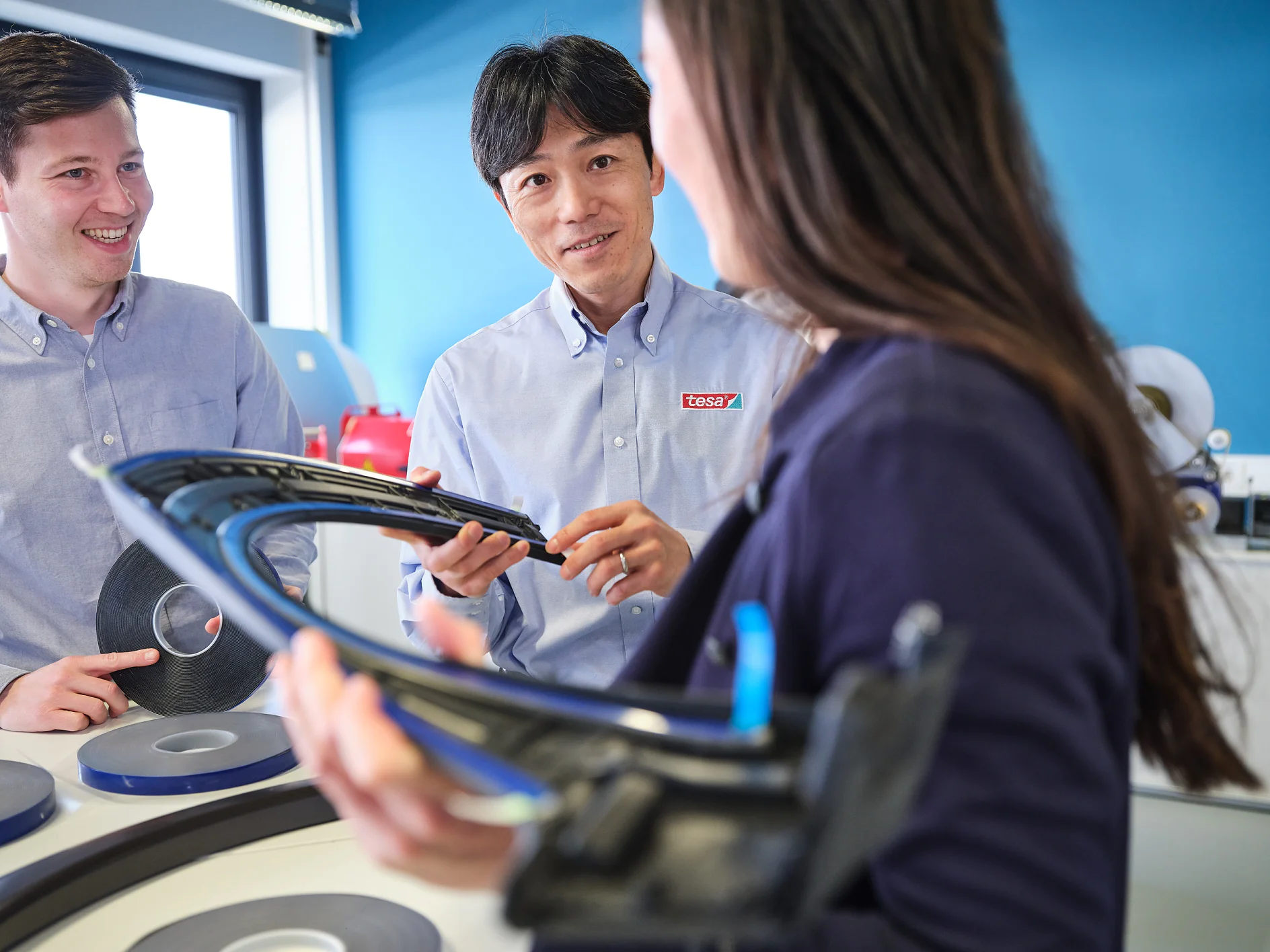 tesa-automotive-key-visual-exterior-005-300dpi Three people in a workspace examine a curved automotive part. Two men in light blue shirts converse with a woman in a dark blazer. Rolls of tesa tape are on the table nearby. The background features a blue wall and a window. (This text has been generated by AI)