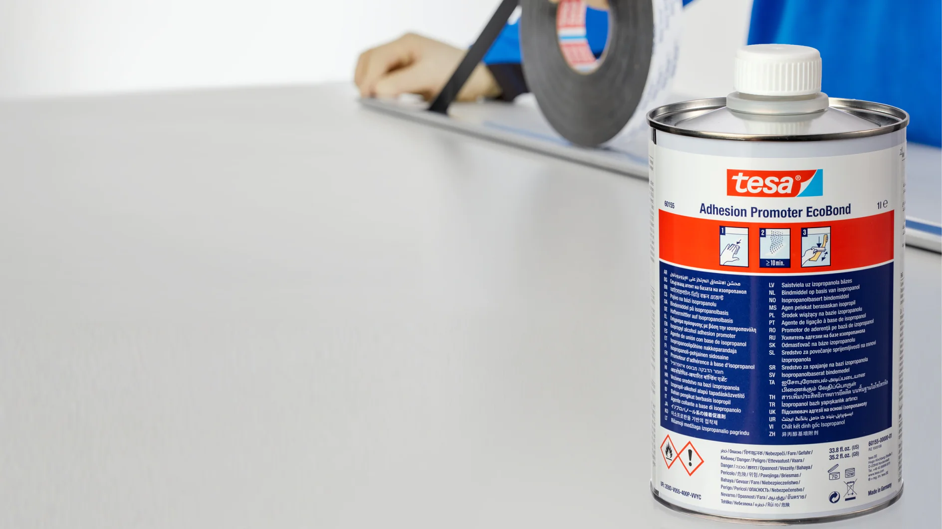 A person in a blue shirt wearing gloves holds a roll of tesa tape above a flat white surface. In the foreground, a can of tesa Adhesion Promoter EcoBond is visible with text and hazard symbols on its label. (This text has been generated by AI)
