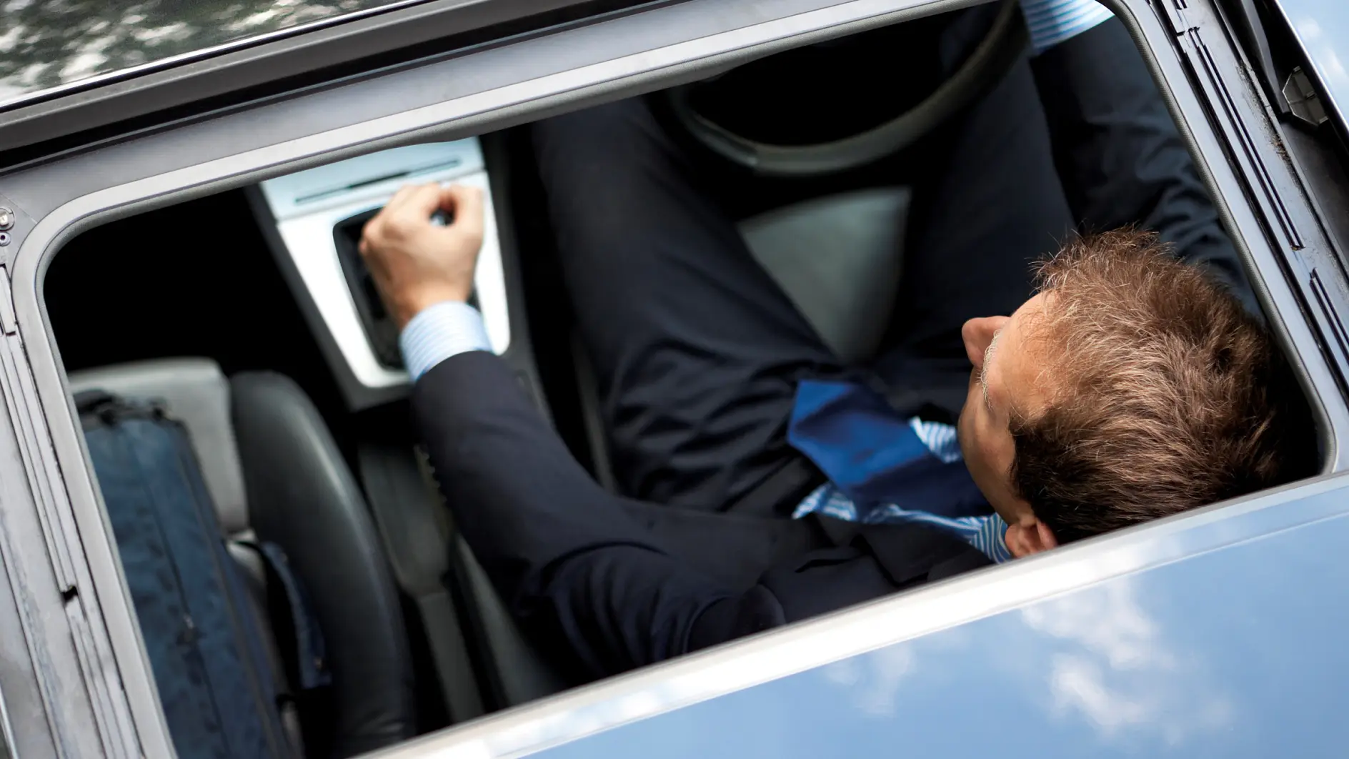 A man in a suit sits inside a car, viewed through an open sunroof, adjusting dashboard controls in a modern interior—enhanced with reliable Automotive tape solutions.