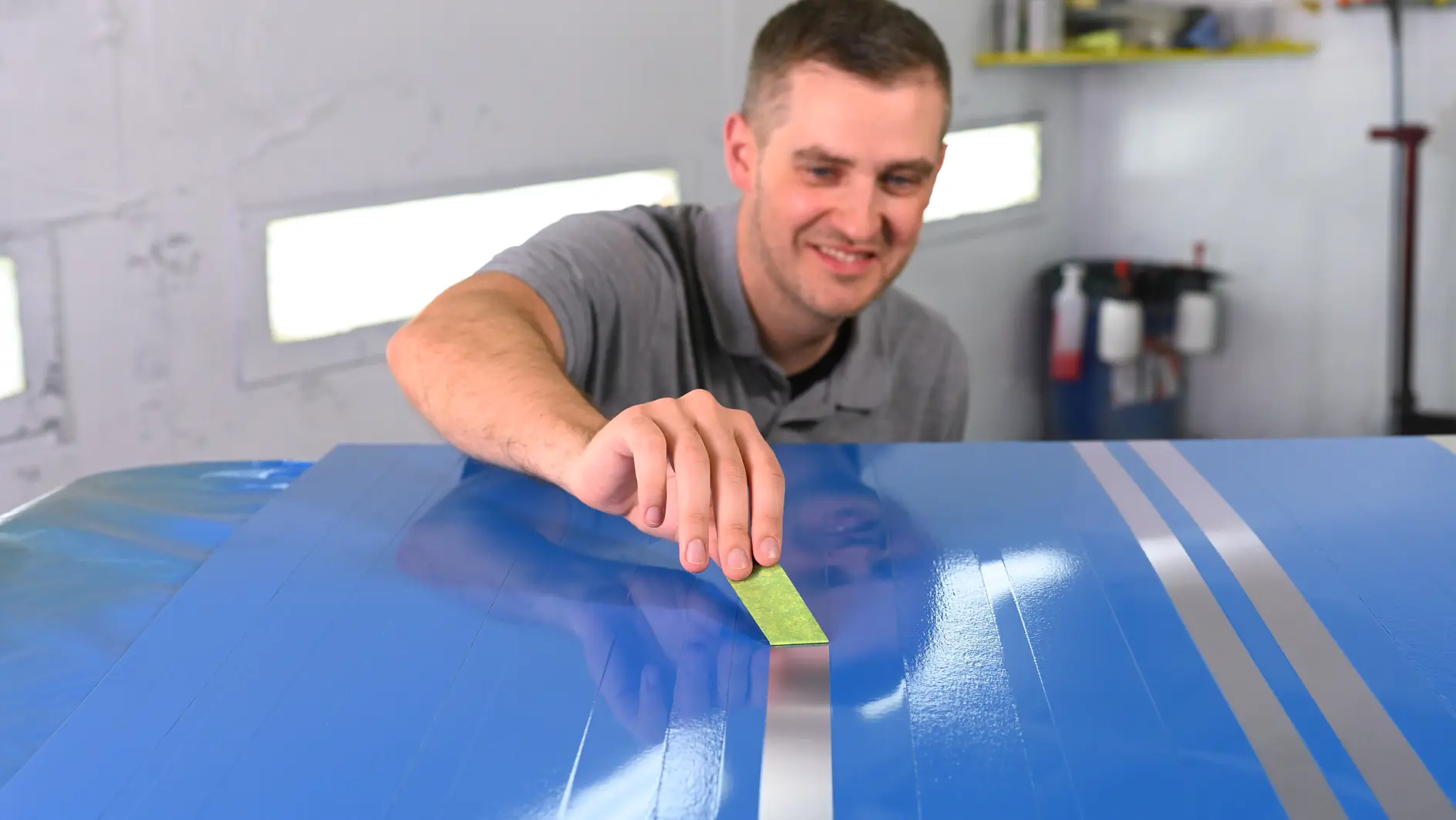 A person in a gray shirt applies a green strip of tesa tape to a blue surface with light gray stripes in a workshop. Shelves and equipment are visible in the background.