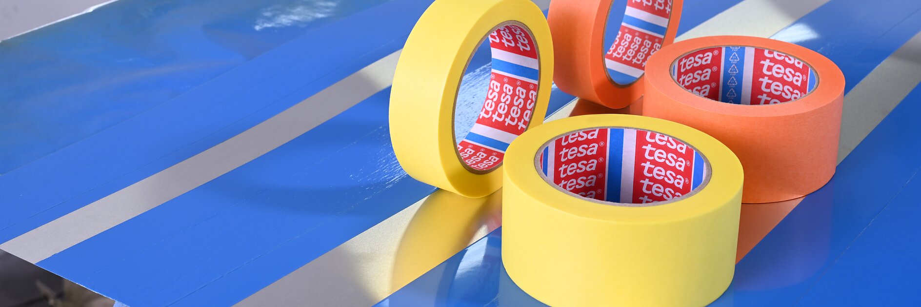 Four rolls of tesa tape—two yellow, one orange, and one bright orange—are positioned on a blue surface. The surface has silver lines, and the background includes a window and blurred objects.