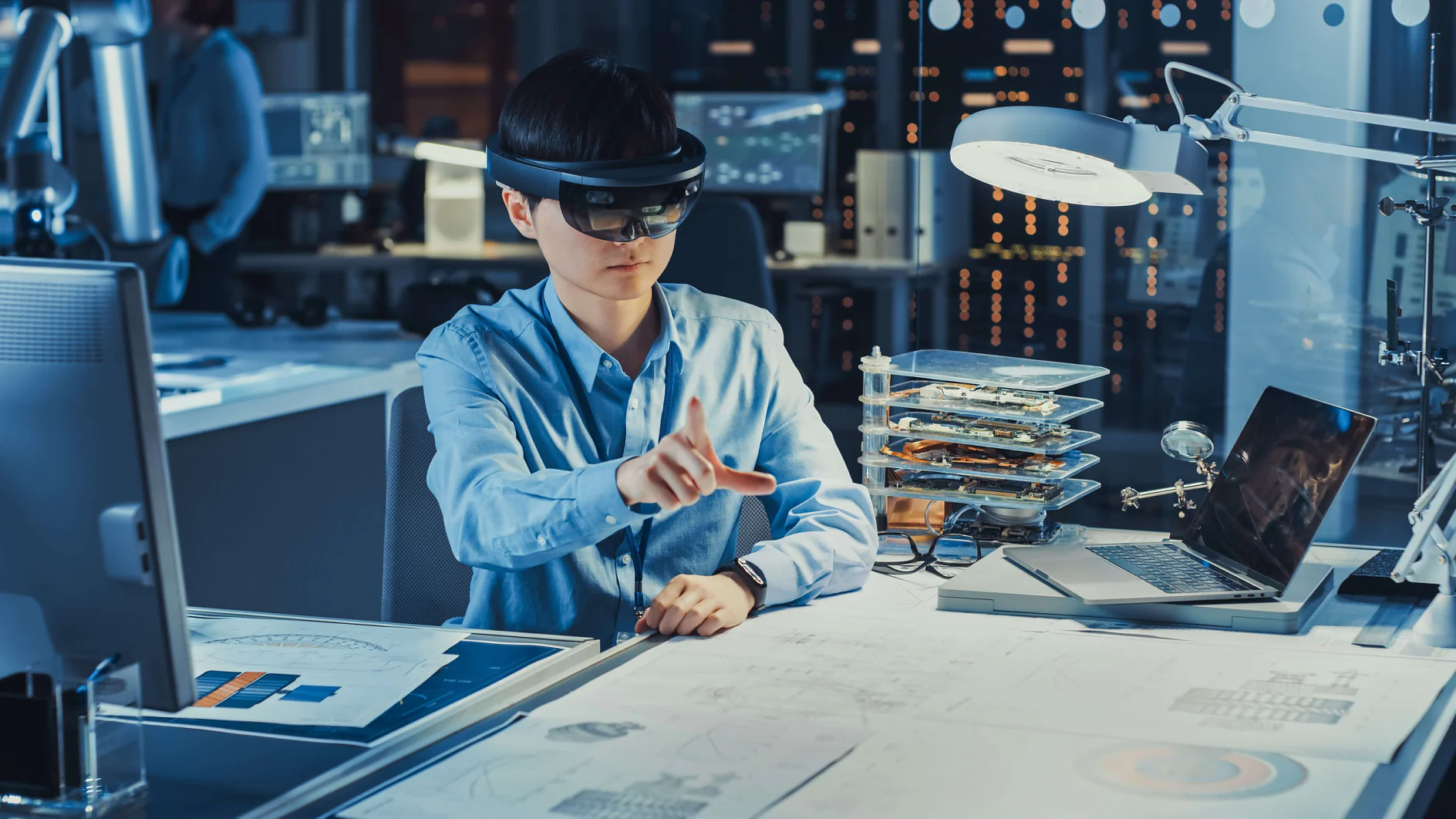 A person wearing an augmented reality headset works at a desk with blueprints, a laptop, and a stack of small electronic devices. The environment appears to be a modern office or lab with digital displays in the background. Various items on the desk are securely fastened using tesa tape. (This text has been generated by AI)