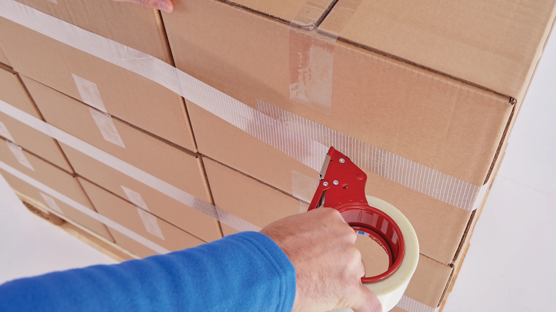 hand in blue sleeve sealing stacked cardboard boxes with clear tesa tape in indoor setting (This text has been generated by AI)