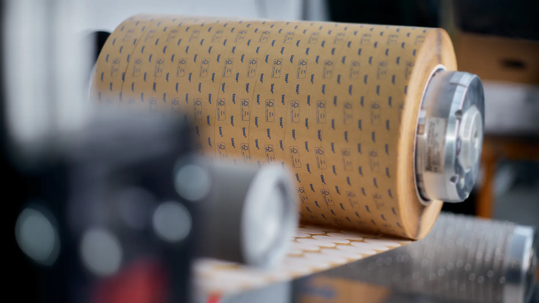 Close-up of a large roll of tesa tape mounted on a metal spindle in a manufacturing environment, highlighting compostable tape solutions for sustainable industrial packaging and production processes.