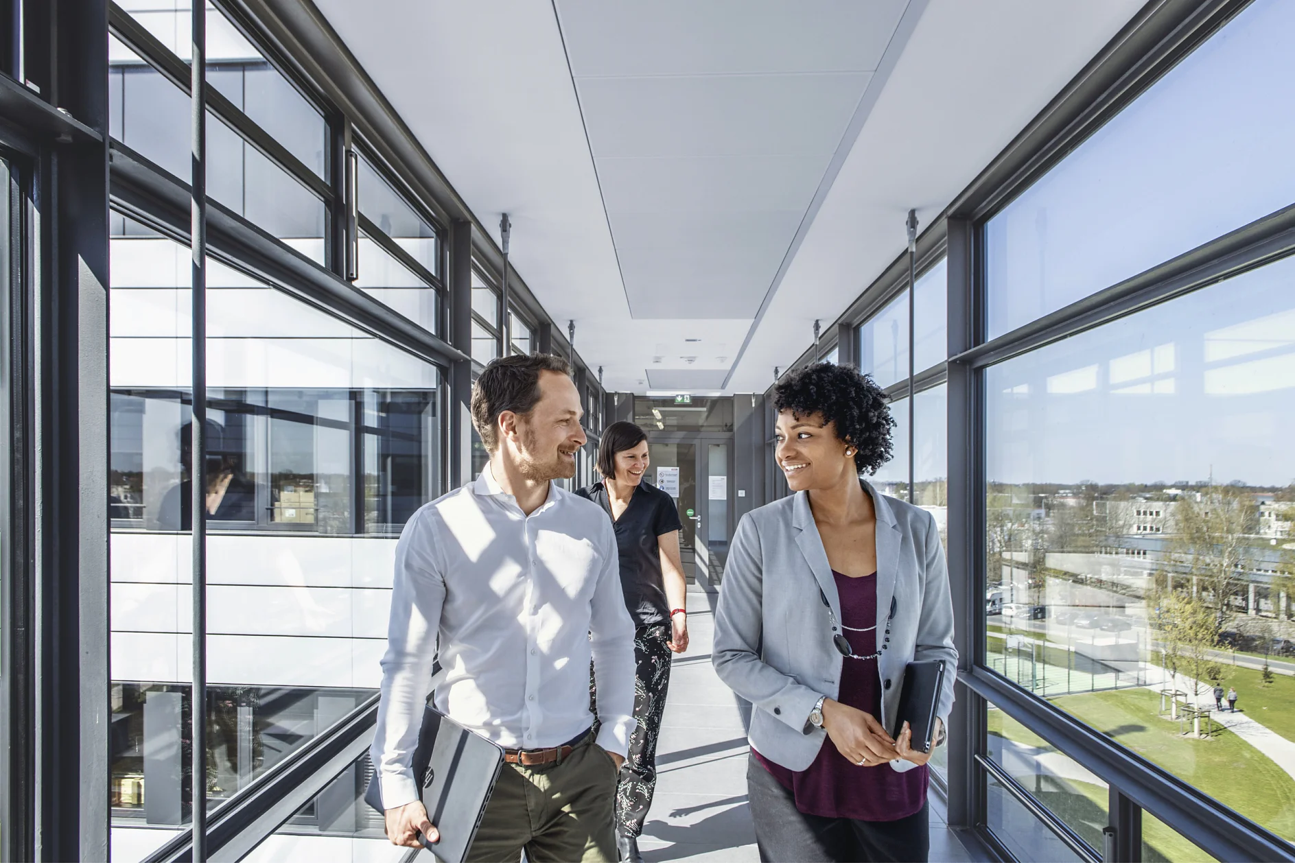 Two people are walking down a bright, modern corridor with large windows, holding tesa tape. They appear to be engaged in conversation. A third person is walking behind them. The outside view shows a clear day and buildings. (This text has been generated by AI)