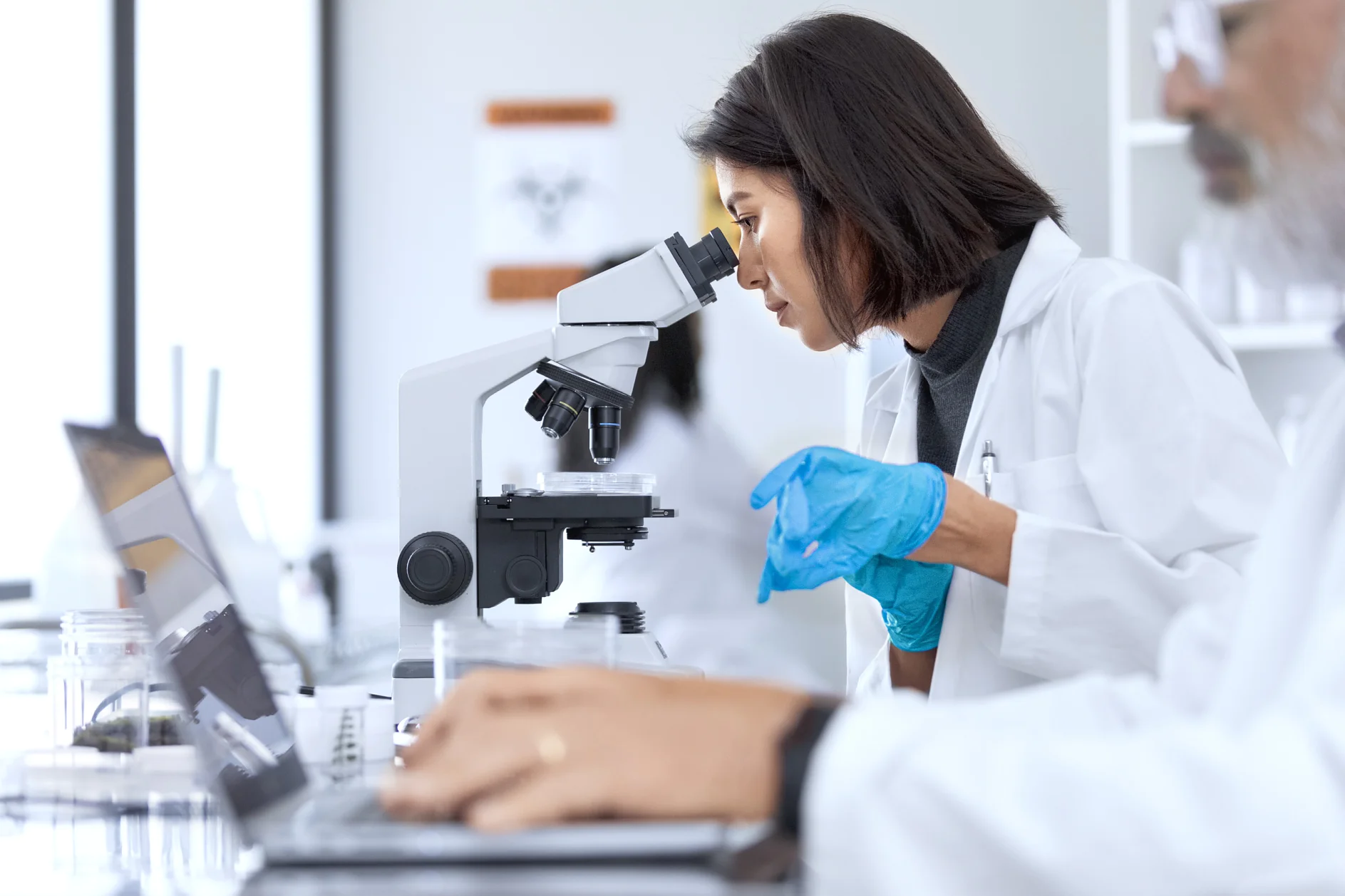 A woman in a lab coat uses a microscope in a laboratory setting. She wears blue gloves and focuses intently. In the foreground, a man in a lab coat works on a laptop. Scientific equipment, including tesa tape, is visible around them. (This text has been generated by AI)