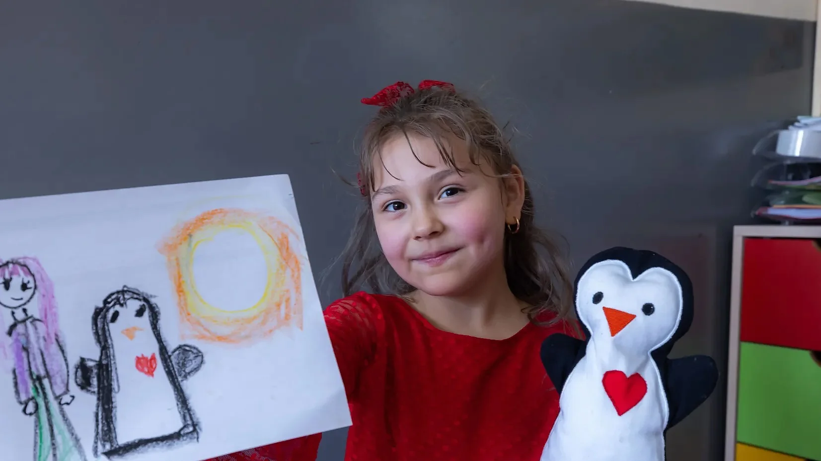 A young girl in a red dress sits at a red table, holding up a drawing of a person, a penguin, and a sun that has been affixed using tesa tape, while also holding a penguin hand puppet with a red heart on its chest. (This text has been generated by AI)