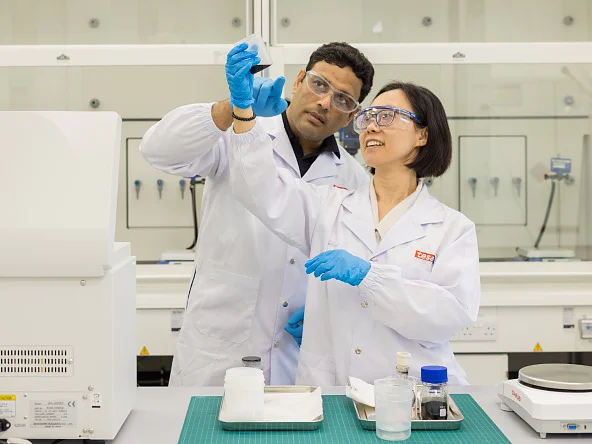 Two scientists in lab coats, safety glasses, and gloves examine a sample in a lab with tesa tape, equipment, and white cabinets. (This text has been generated by AI)