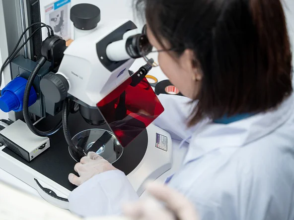 A person in a lab coat and gloves uses a microscope to examine a slide. Lab equipment, tesa tape, and a red filter are nearby. (This text has been generated by AI)