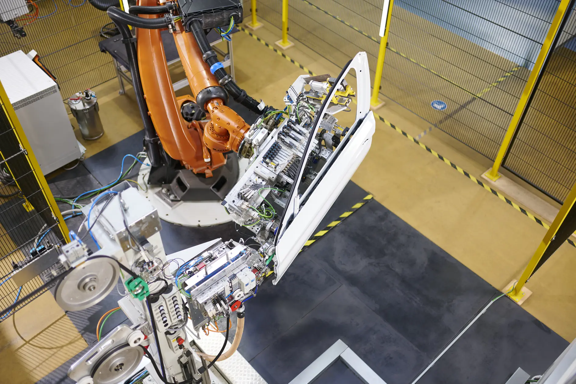 A robotic arm installs a car door using tesa tape in an industrial facility, with yellow safety barriers and equipment in view.