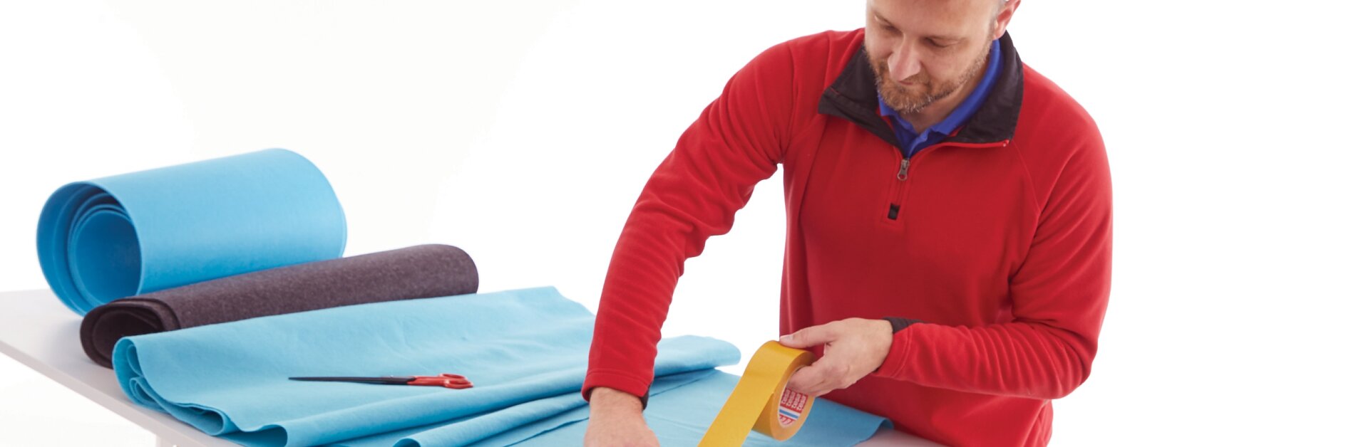 man in red sweater cutting blue foam sheets on white table with rolls of foam and yellow tape indoors