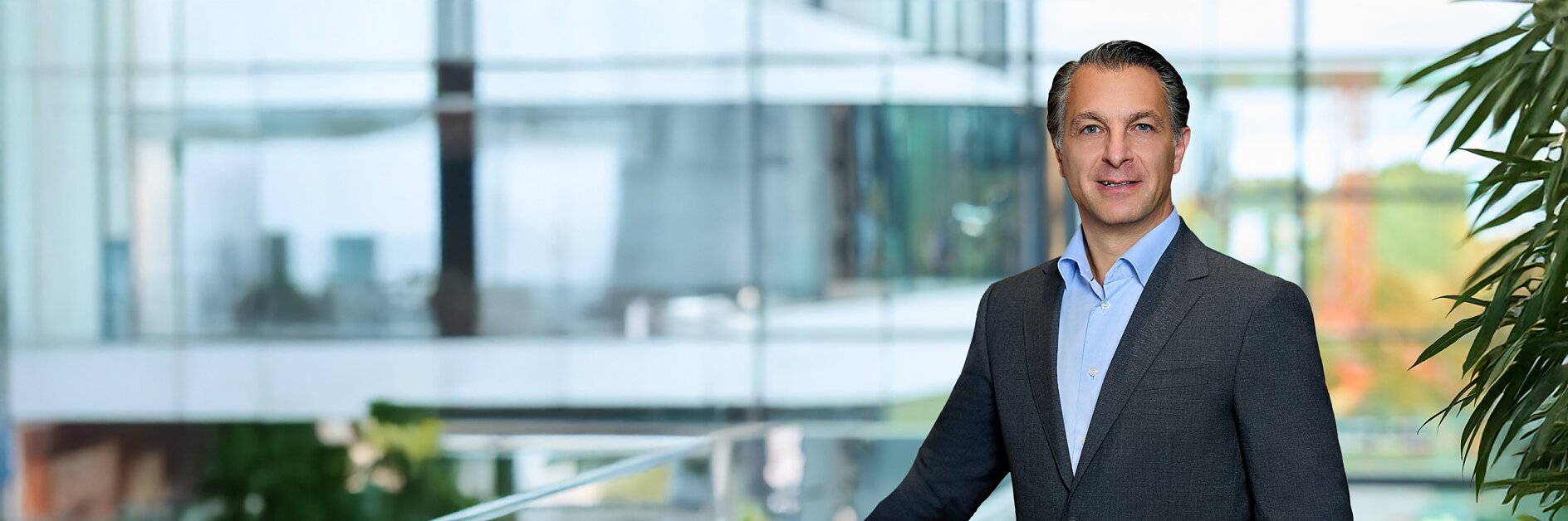 A man in a dark suit and light blue shirt stands by a glass railing indoors, with modern architecture and blurred greenery behind him. (This text has been generated by AI)