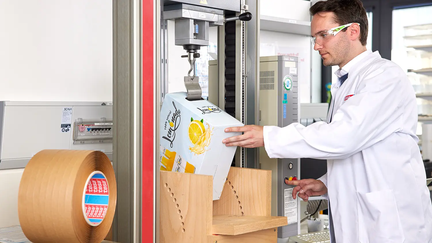 A man in a white lab coat and safety glasses operates a packaging tester near a roll of tesa tape and other lab equipment, evaluating the performance and durability of recyclable tape solutions in a laboratory environment.
