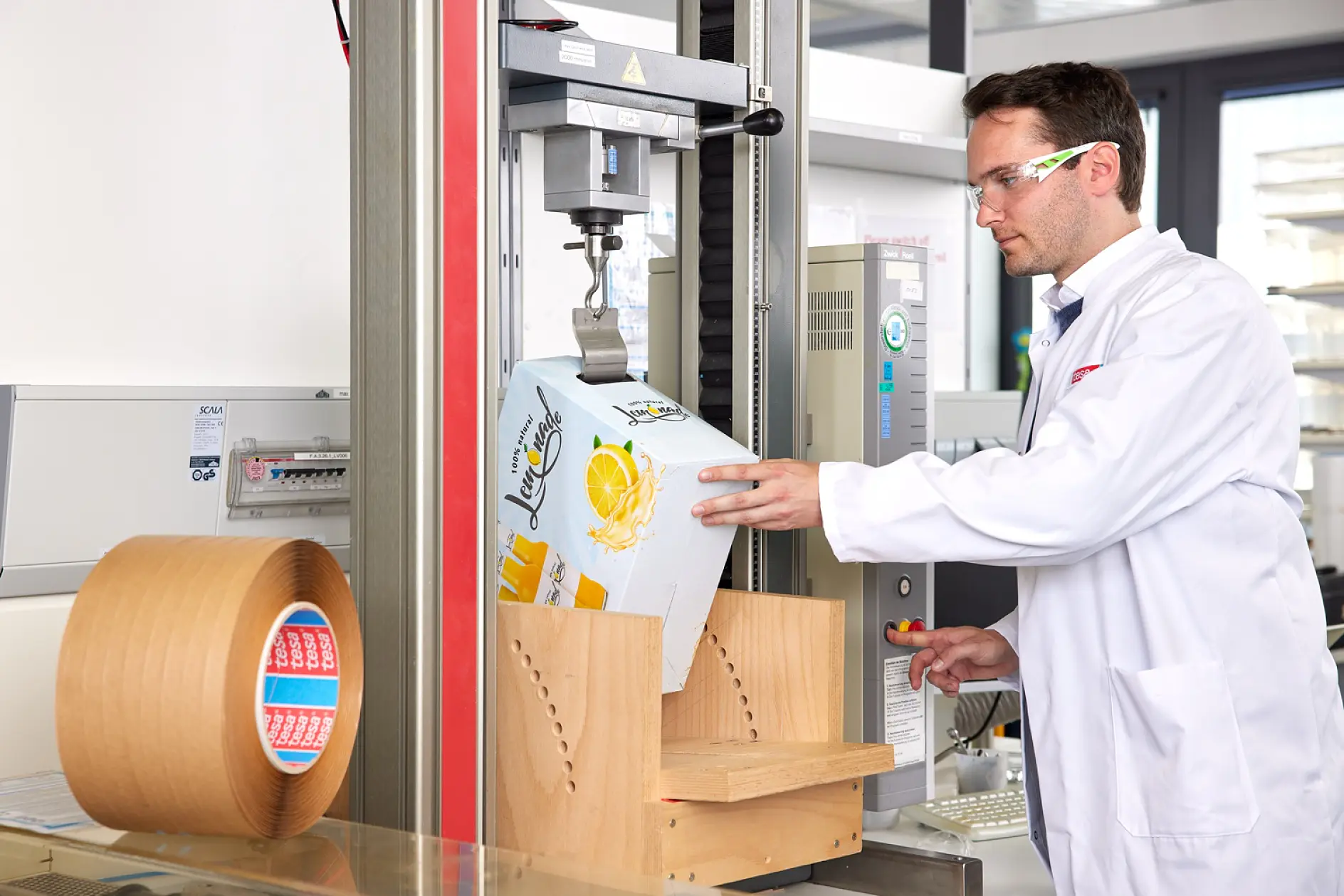A man in a white lab coat and safety glasses operates a packaging tester near a roll of tesa tape and other lab equipment, evaluating the performance and durability of recyclable tape solutions in a laboratory environment.