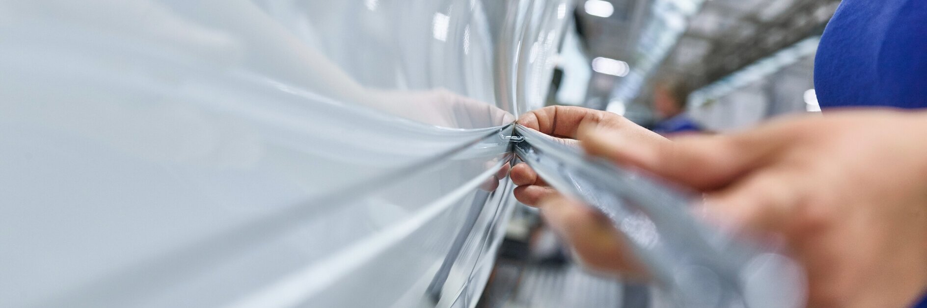 tesa tape being applied to a white vehicle in a factory setting, focusing on hands smoothing the tape along the surface