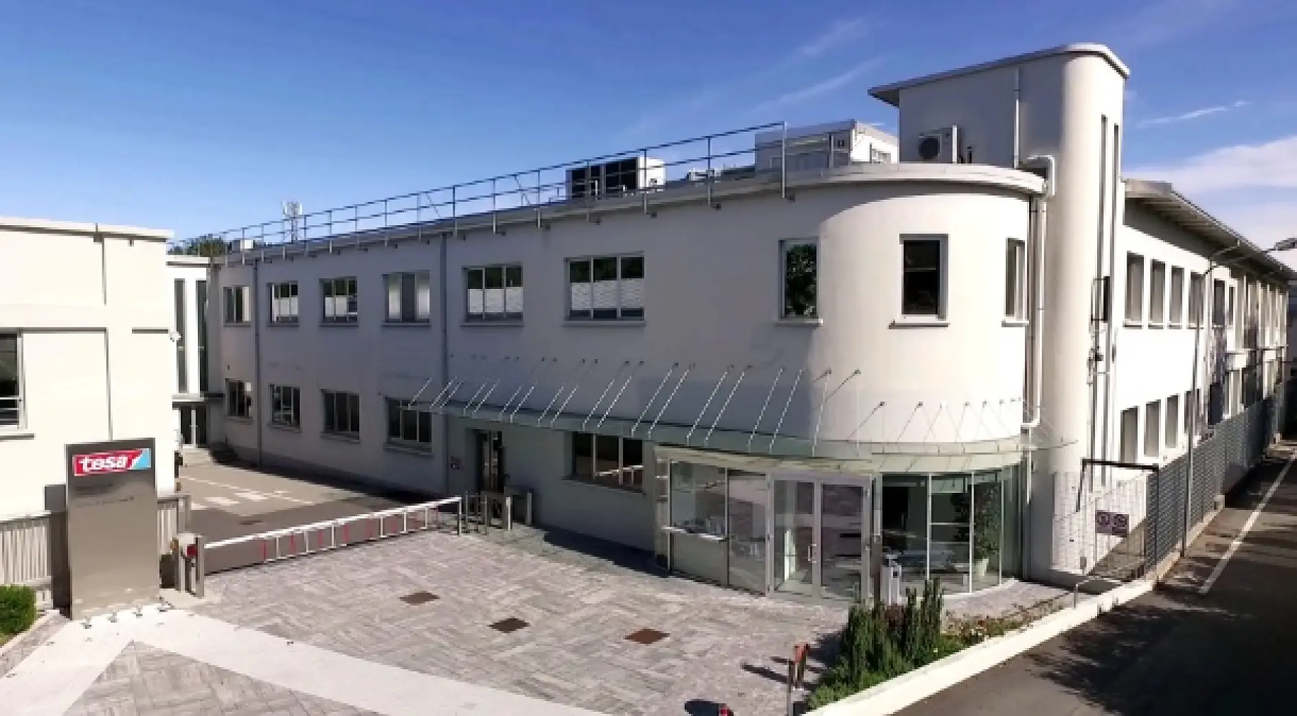 A modern white two-story office with many windows and a “tesa tape” sign near the glass entrance, photographed on a sunny day. (This text has been generated by AI)