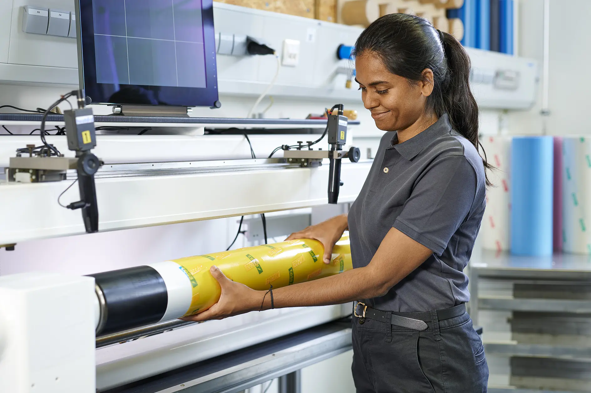 woman in gray tesa shirt applying yellow adhesive film on black cylindrical object in bright workshop with computer and tools (This text has been generated by AI)