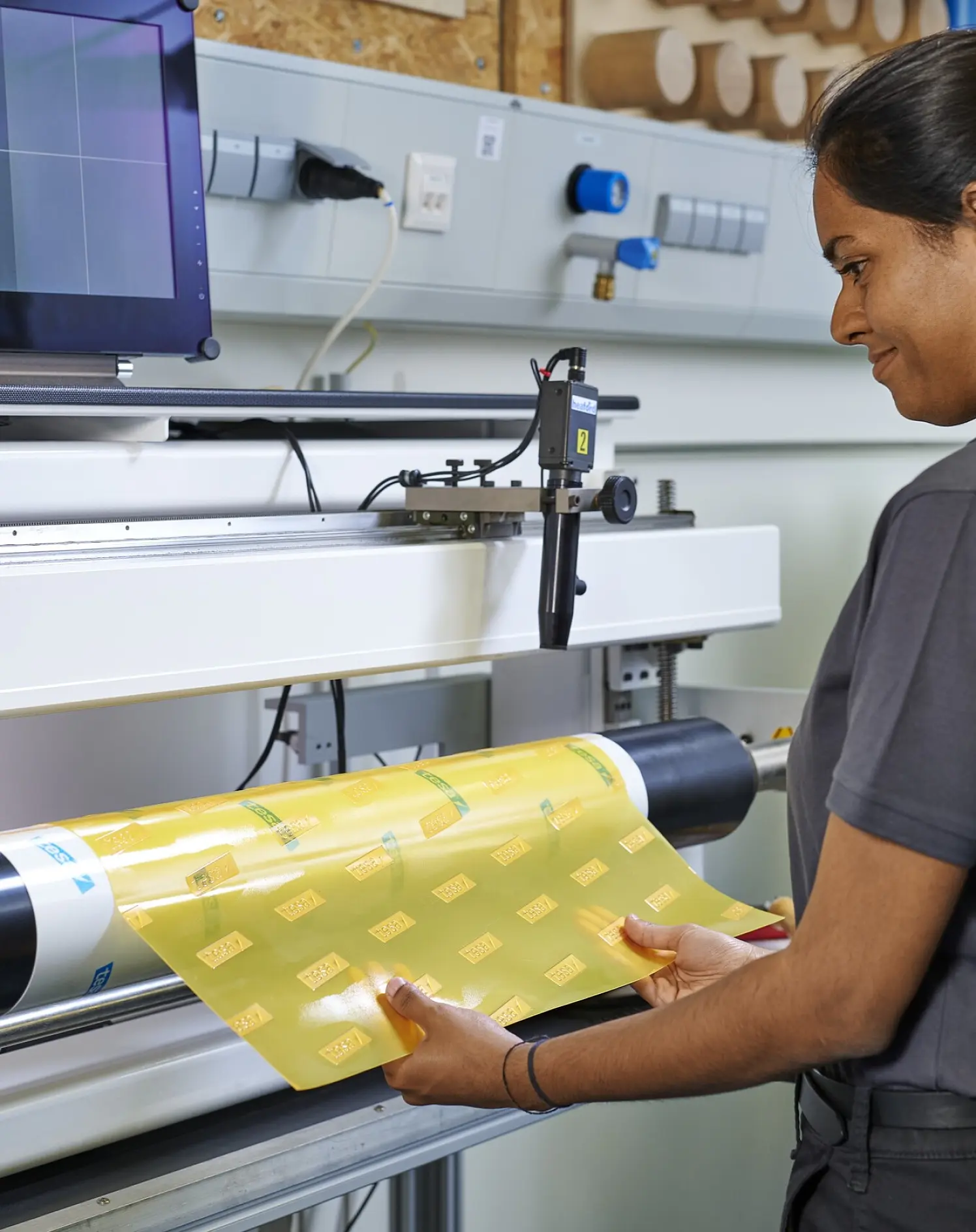 A woman operates a machine processing yellow sheets of repeating tesa tape labels, holding one as it feeds past a monitor and controls. (This text has been generated by AI)
