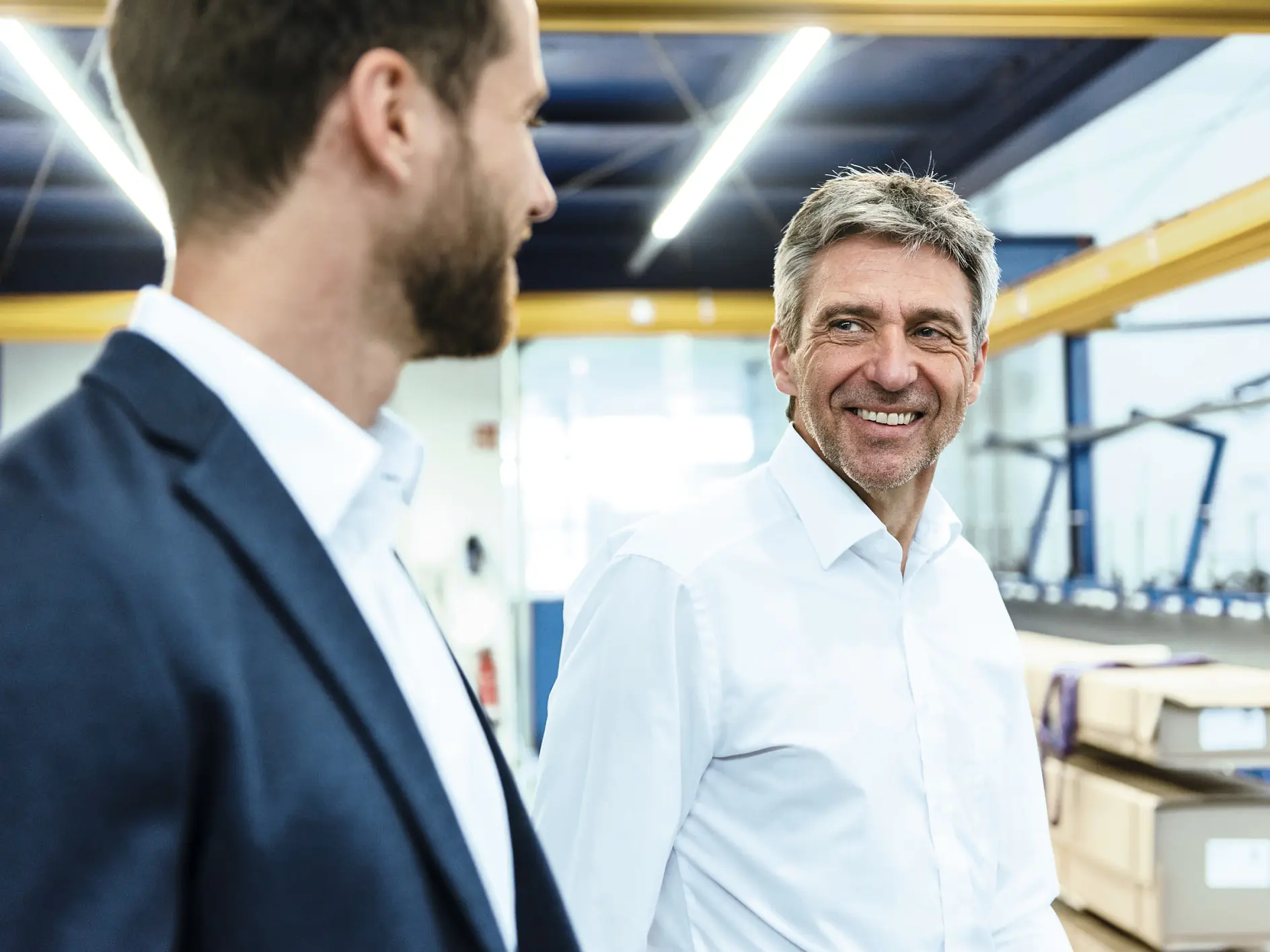 Two men in business attire discuss inside a warehouse with shelves and stacked boxes in the background. (This text has been generated by AI)