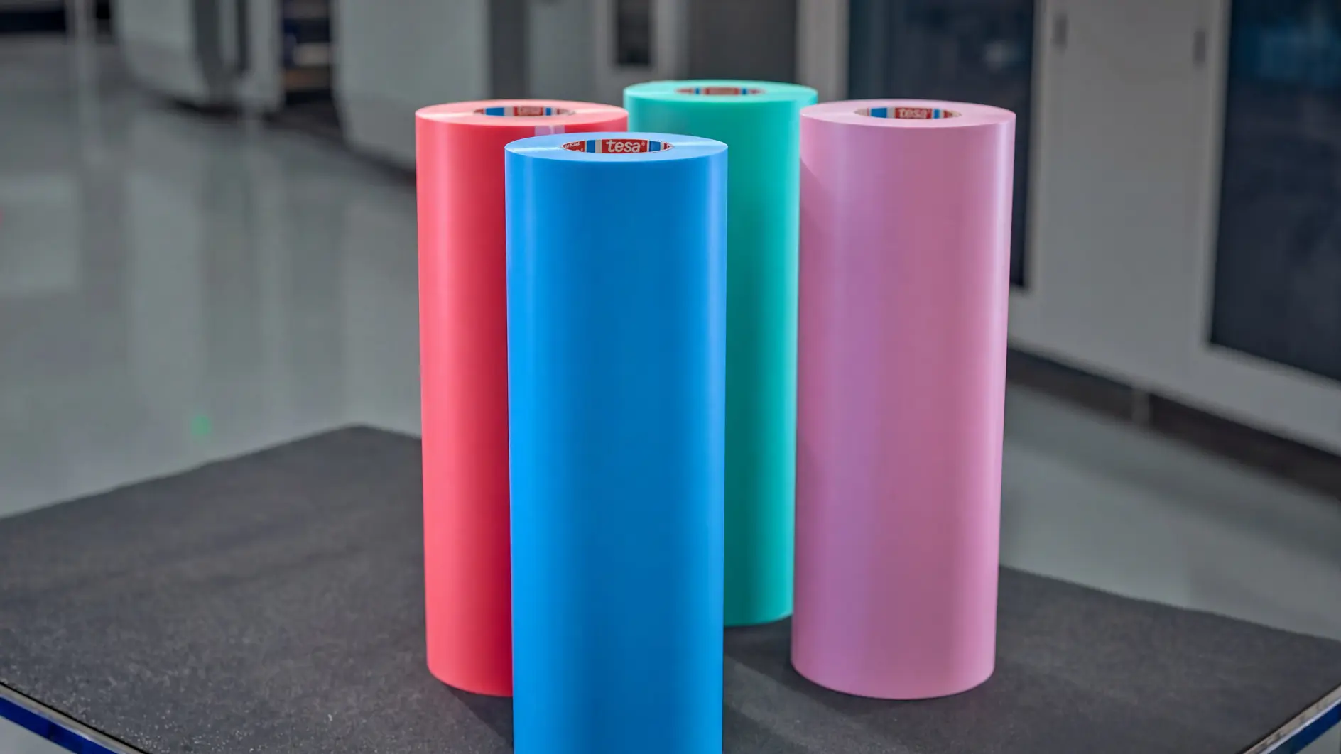 Five colorful rolls of tesa adhesive film standing upright on a black surface in an indoor industrial setting. (This text has been generated by AI)