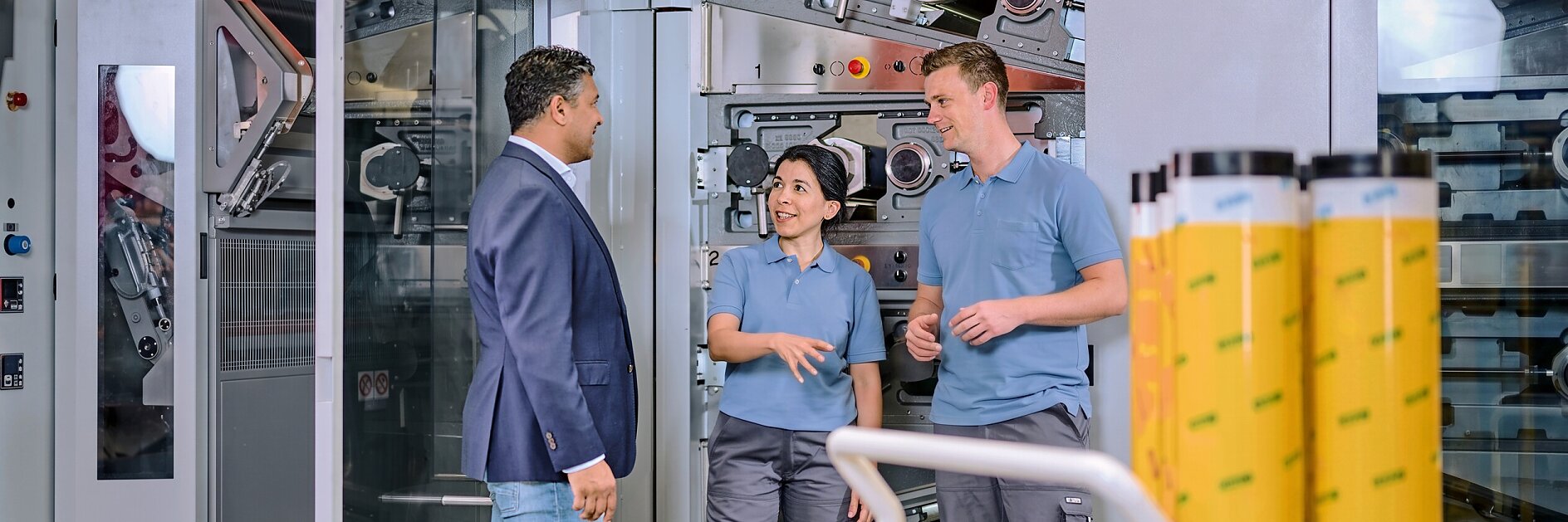 Three people in a factory setting examine large rolls of yellow tesa adhesive tape, surrounded by industrial machinery and equipment.