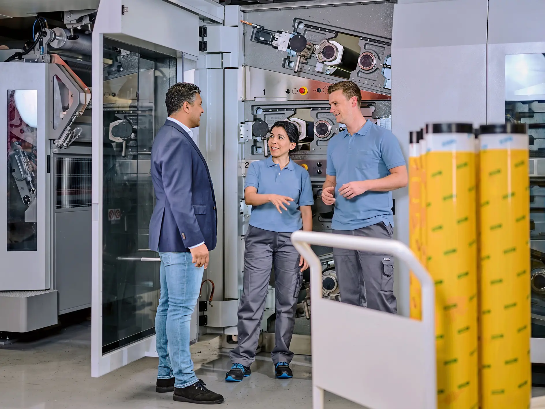 Three people in a factory setting examine large rolls of yellow tesa adhesive tape, surrounded by industrial machinery and equipment. (This text has been generated by AI)