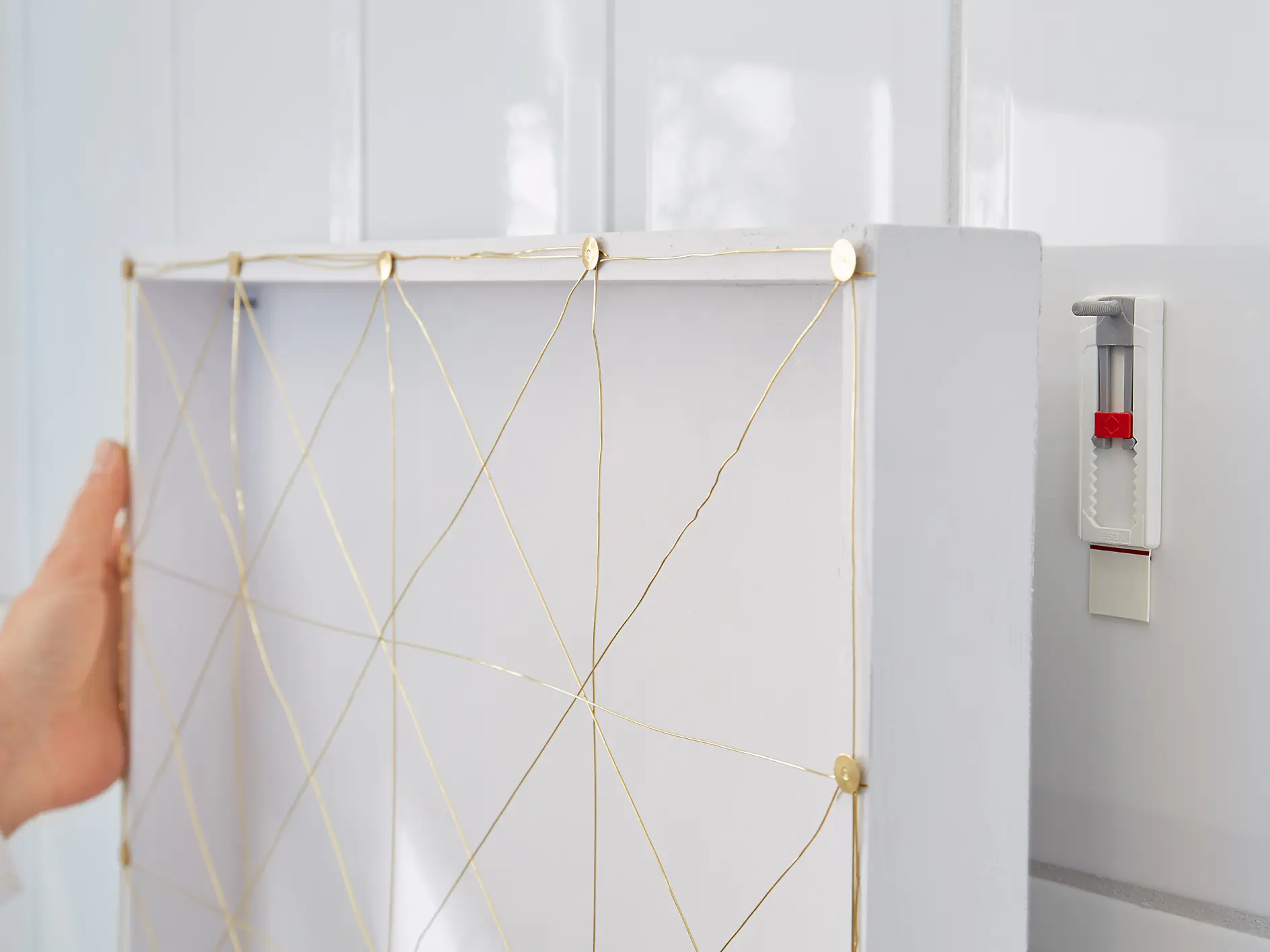 A person is hanging a white photo frame on a white tiled wall using a small level tool attached to the wall with tesa tape. The frame has a crisscross pattern made of gold-colored strings.