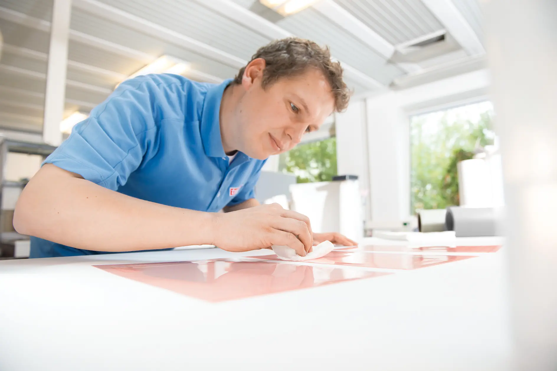 A person in a blue shirt is working intently on a large sheet of paper or material, using a white cloth and tesa tape. The setting is a well-lit room with bright windows and visible greenery outside.