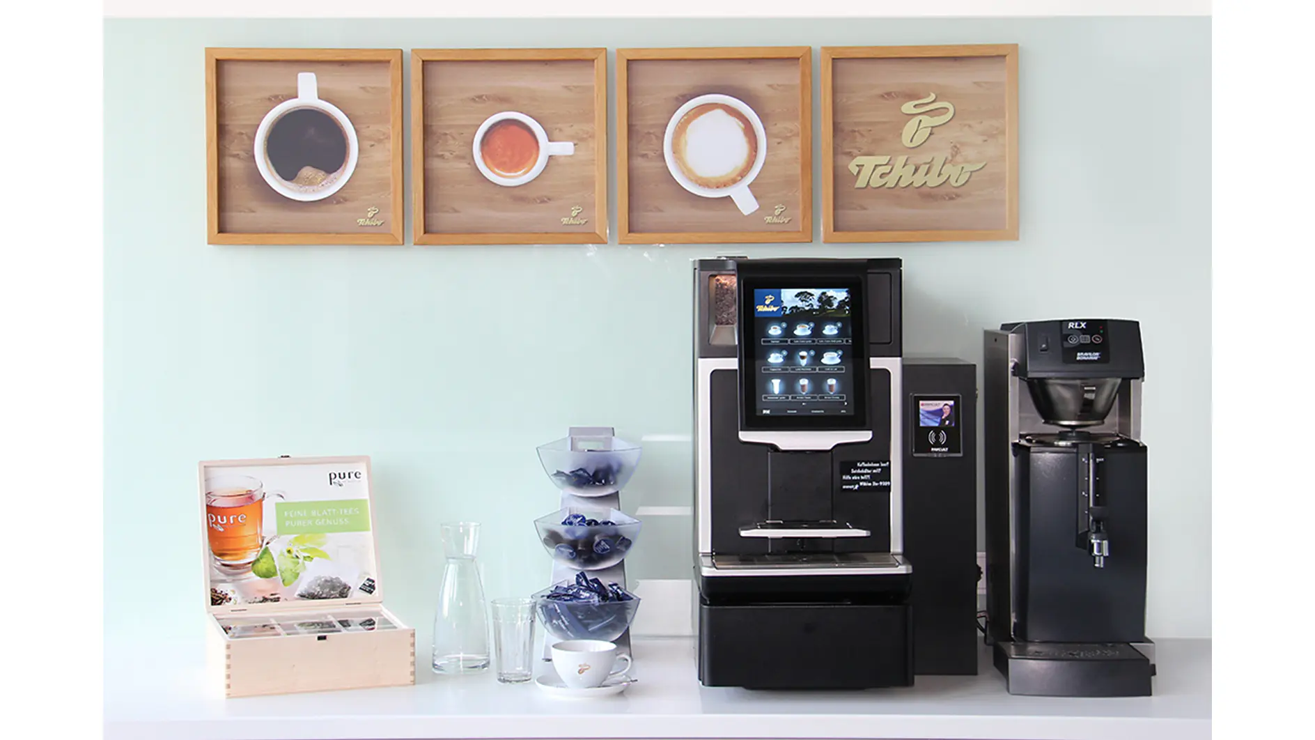 A coffee station featuring two coffee machines on a white countertop. Above them are three framed images of different coffee styles. To the left, there are tesa tape packets, a glass carafe, three tiered bowls with capsules, and a cup. (This text has been generated by AI)