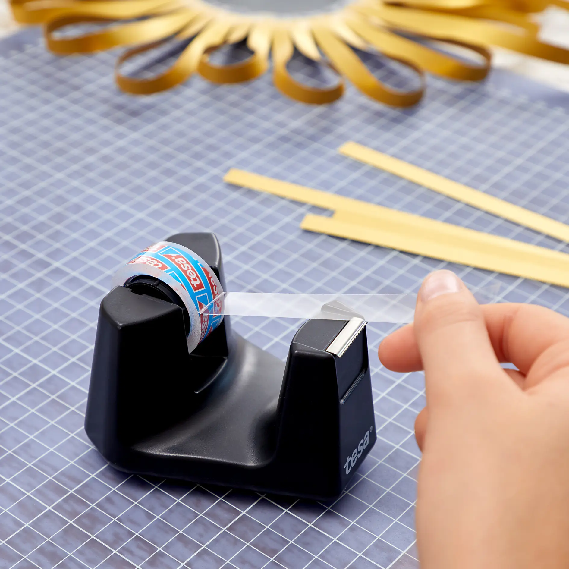 A hand pulls clear tesa tape from a black tape dispenser marked tesa on a checkered cutting mat. Yellow strips and a decorative paper design are visible in the background.