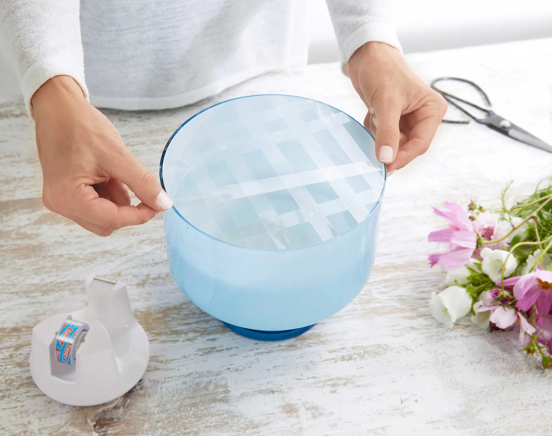 A person is placing a grid of clear tesa tape over the top of a blue vase on a wooden surface. Nearby, there is a small bouquet of pink and white flowers, a pair of scissors, and a tesa tape dispenser.