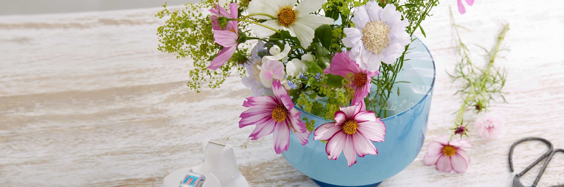 A bouquet of pink, white, and green flowers in a blue glass vase sits on a rustic wooden table. Nearby are a roll of clear tesa tape and a pair of scissors. The background is softly blurred.