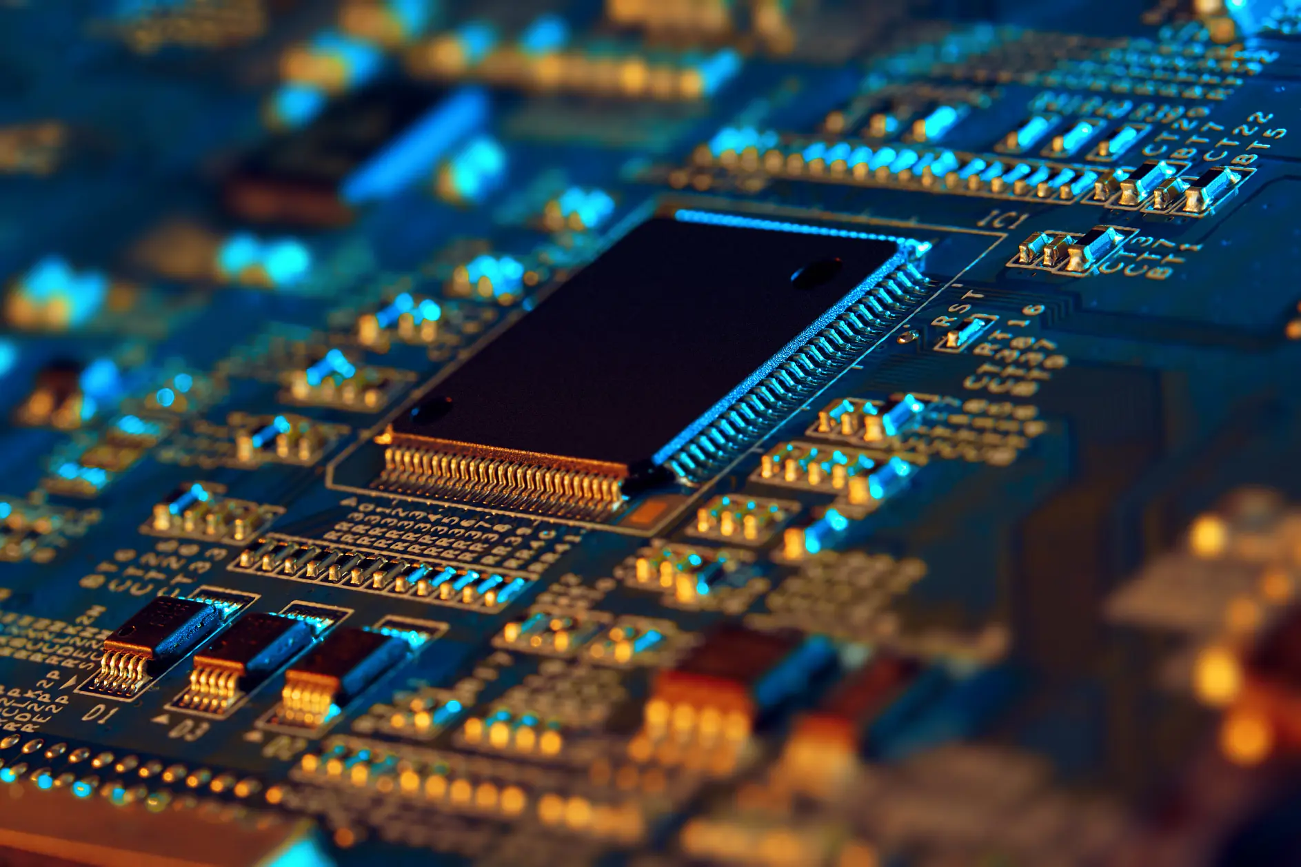 Close-up view of a computer circuit board featuring a central black microchip surrounded by various electronic components and pathways. Blue and orange lighting highlights the intricate details and connections on the board, emphasizing the use of tesa tape in securing certain components.