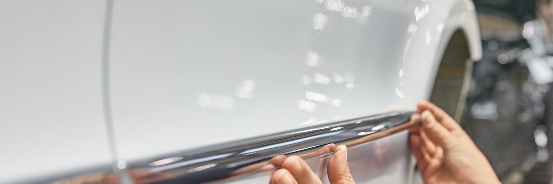 Hands are attaching a chrome trim strip along the side of a shiny white car using tesa tape. The background shows an indoor setting with blurred equipment, indicating an automotive workshop or garage.