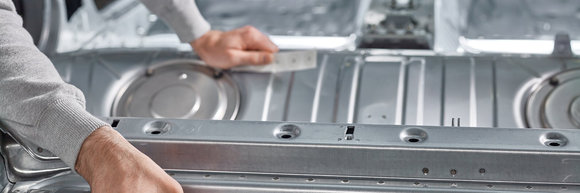 A person is assembling or inspecting a metallic automotive component. Their hands are adjusting or examining sections in the interior body frame of a car, using tesa tape. The background reveals a spacious, unpainted vehicle interior focused on the metal structure.