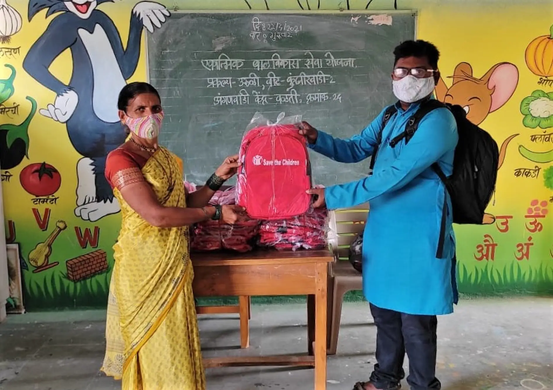 Two people wearing face masks are exchanging a red backpack in a colorful classroom. The chalkboard in the background has writing in Hindi. A stack of similar backpacks is on a table between them, held together with tesa tape. The walls have cartoon character illustrations.