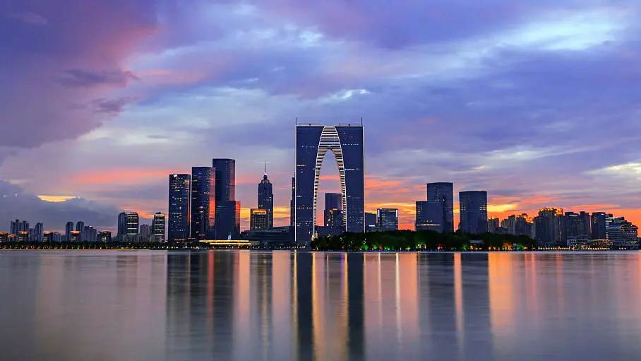 City skyline at sunset, featuring modern skyscrapers and a distinct, large arch-shaped building reflected in calm water. The sky has hues of purple, pink, and orange, creating a vibrant backdrop for the urban landscape.