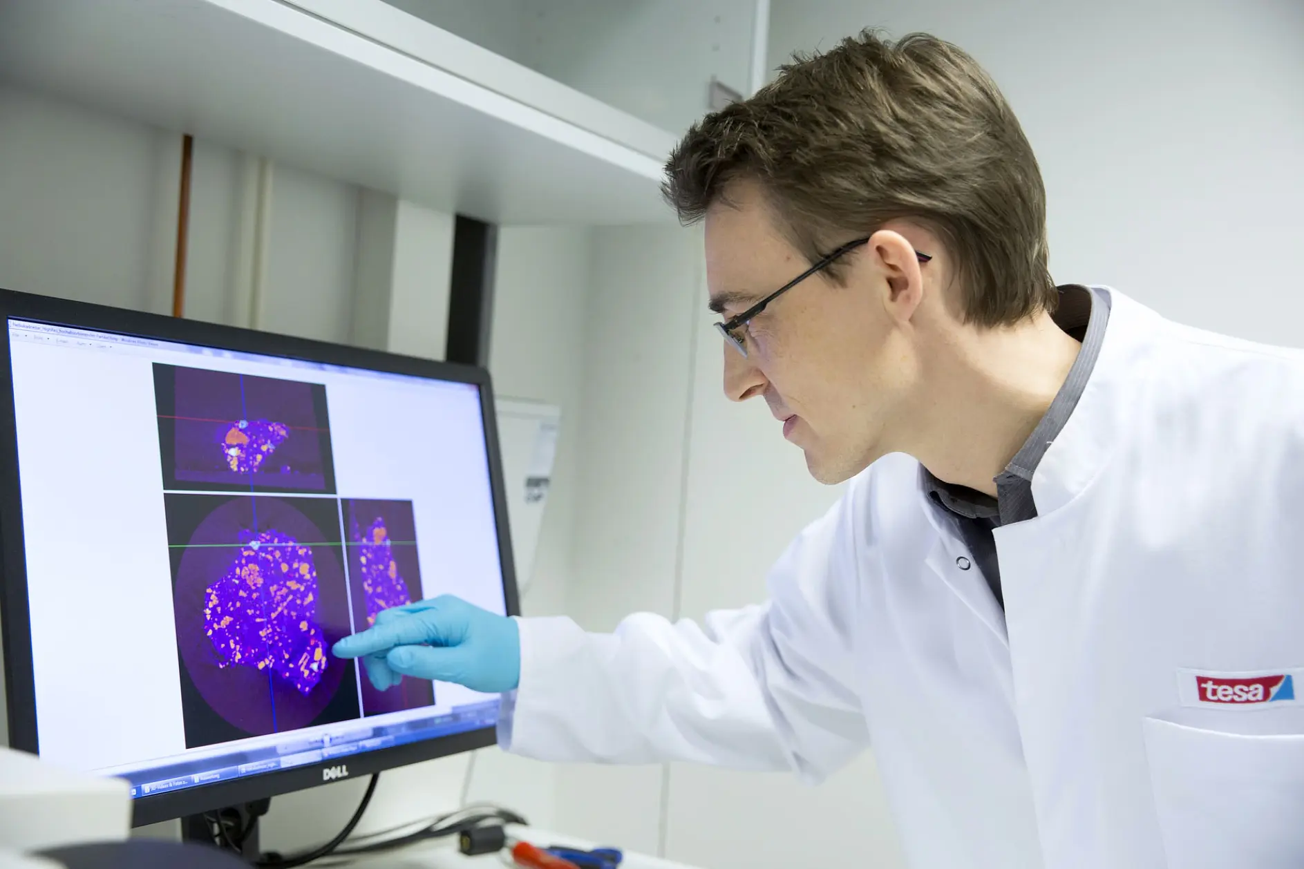 A person in a lab coat and blue gloves examines a computer screen displaying scientific images with purple patterns. The individual points at the screen, indicating interest or analysis. The setting appears to be a laboratory, where various supplies, including tesa tape, are likely used for research purposes.