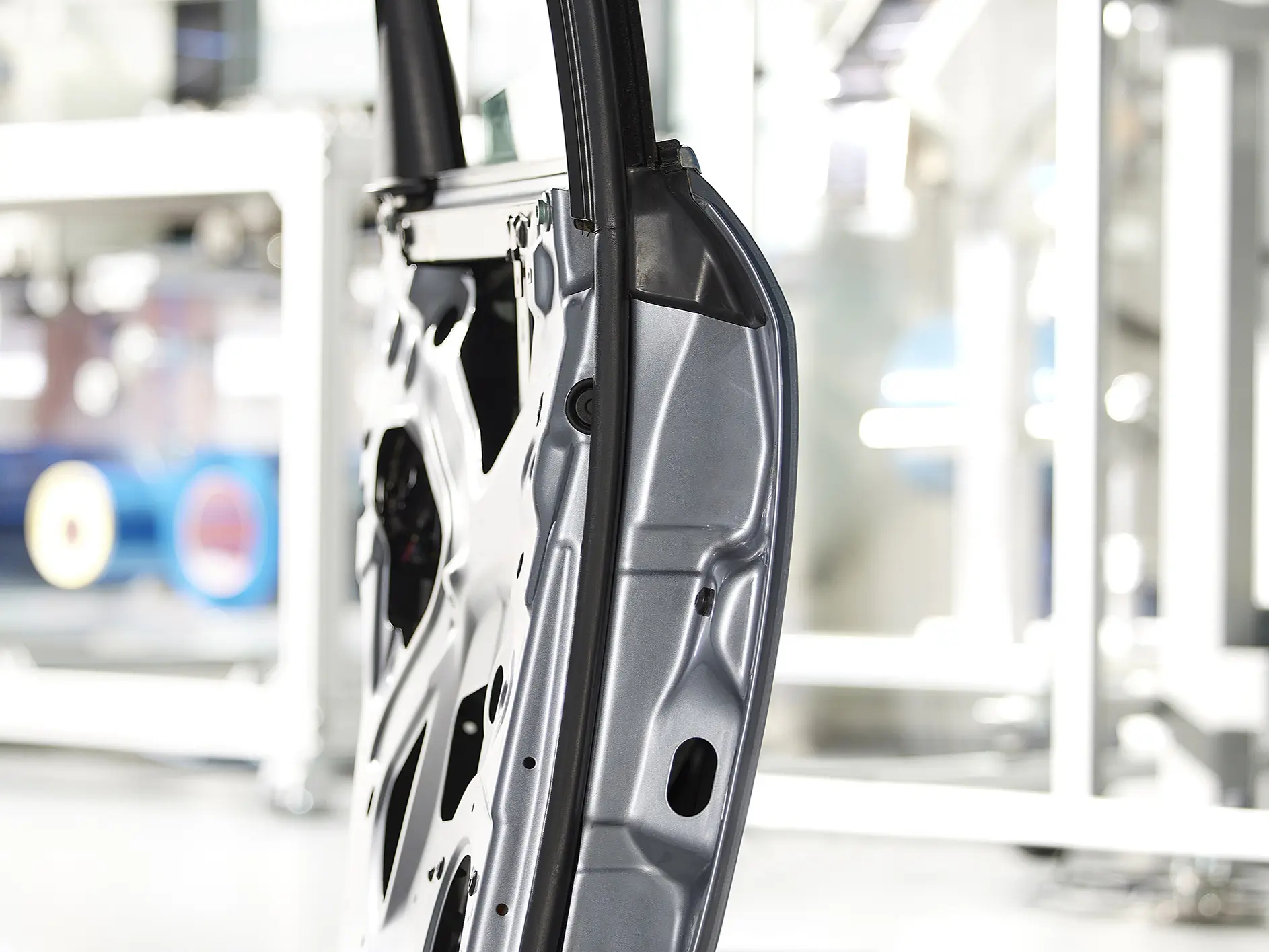 Side view of a metal car door frame in a manufacturing setting. The frame is silver with visible cutouts and structural details, secured with tesa tape. The background is blurred, featuring industrial equipment and machinery. (This text has been generated by AI)
