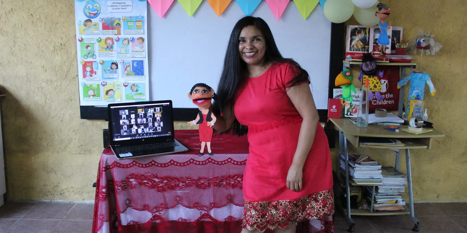 A woman in a red dress stands smiling in a classroom setting. She holds a puppet. A laptop with multiple video call screens is on a red table with a decorative cloth. Colorful banners and educational posters, held up by tesa tape, are in the background.