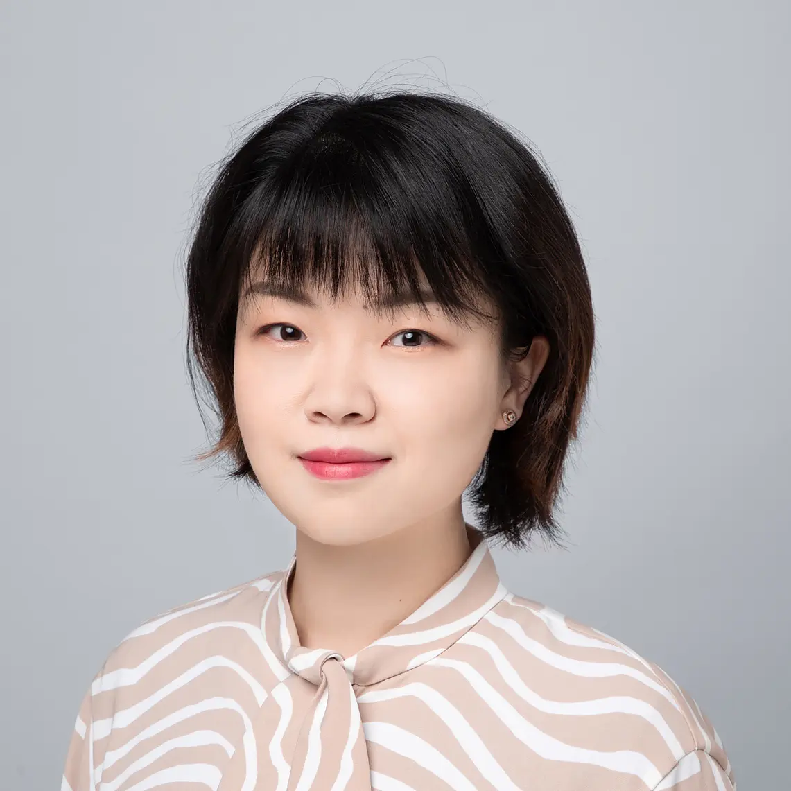 A person with short dark hair and bangs is wearing a blouse with a beige and white wavy pattern. They face the camera against a light gray background.