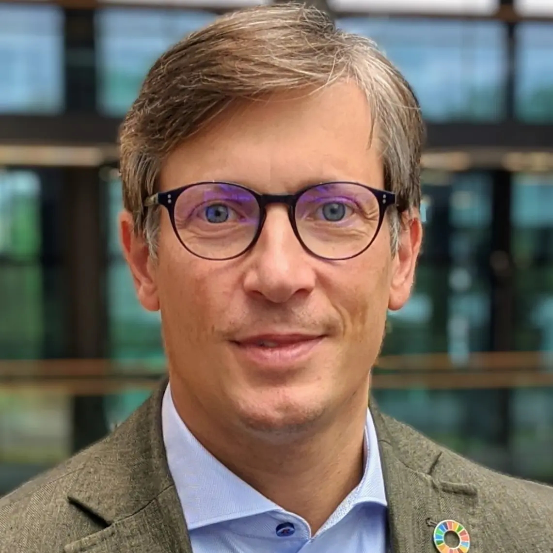 A man with short hair and glasses is wearing a dark green blazer over a light blue shirt. He has a circular pin on his lapel. The background shows an office setting with large windows and green foliage outside.